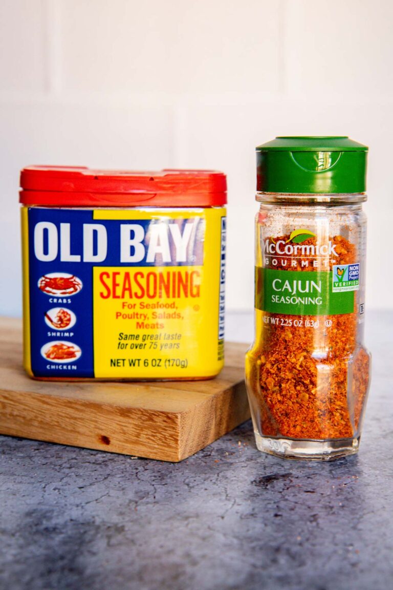 A container of Old Bay seasoning sits next to a bottle of McCormick Gourmet Cajun seasoning on a wooden board, both on a gray countertop—perfect for anyone interested in comparing Old Bay vs Cajun Seasoning in their next recipe.