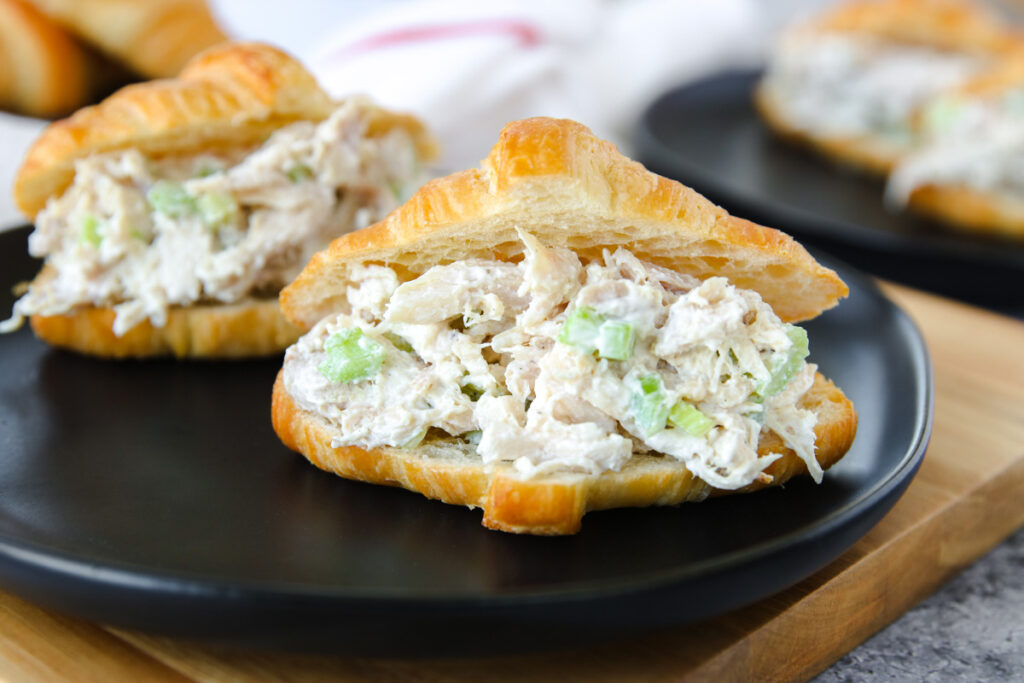A croissant sandwich filled with creamy rotisserie chicken salad and chopped celery, served on a black plate with another sandwich in the background.