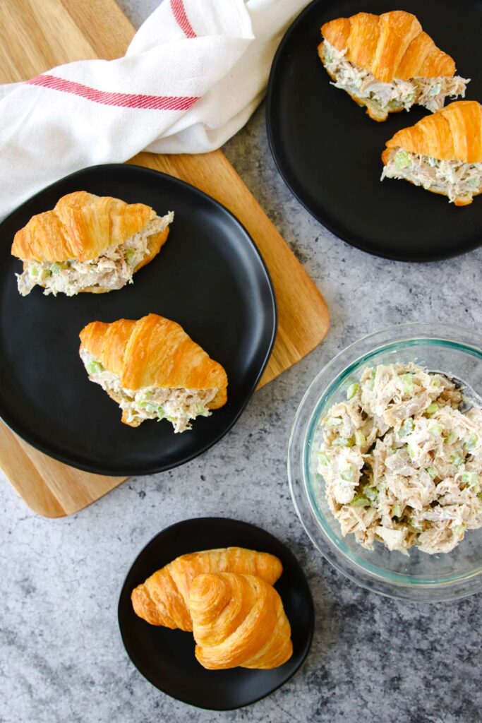 Three small croissant sandwiches filled with creamy rotisserie chicken salad rest on black plates, one halved and one plain croissant beside them. A glass bowl of rotisserie chicken salad and a napkin are also on a gray countertop.