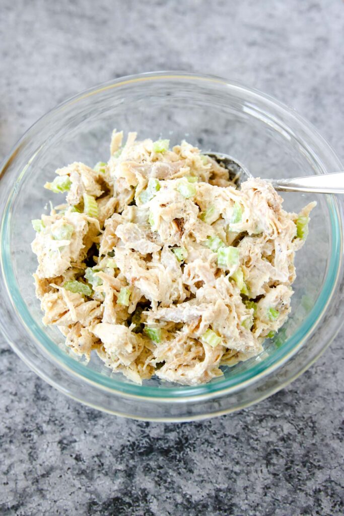 A glass bowl filled with rotisserie chicken salad made with shredded chicken, chopped celery, and mayonnaise, placed on a gray countertop with a spoon inside.