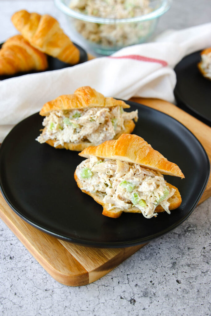Two croissant sandwiches filled with rotisserie chicken salad and celery sit on a black plate. Another croissant and a bowl of rotisserie chicken salad are in the background.